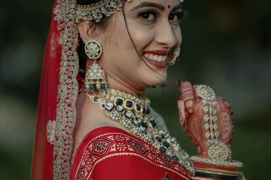 Indian bride in traditional red sari and jewelry smiling outdoors, showcasing cultural elegance.